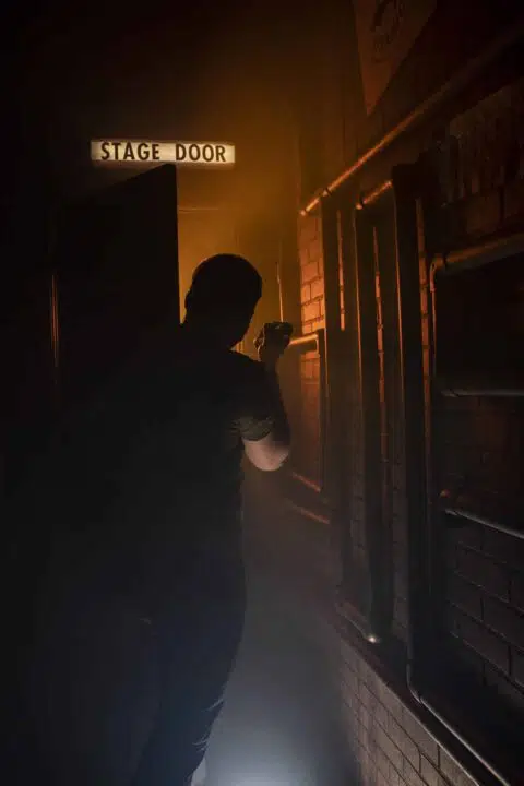 A photograph of a silhouette holding a flashlight in front of the stage door which is glowing orange.